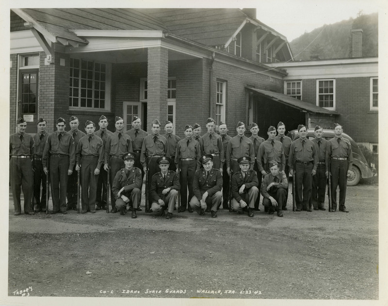 Idaho State Guards members pose for a photo outside in front of a building and an automobile. "CO-L IDAHO STATE GUARDS-WALLACE, IDA  6-23-42" is written on the bottom of the photograph.