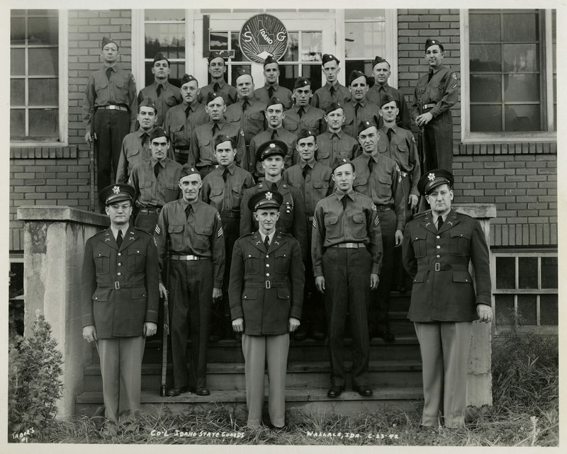 Idaho State Guards members stand together on the steps of a building. "CO-L IDAHO STATE GUARDS WALLACE, IDA. 6-23-74" is written on the bottom of the photograph.
