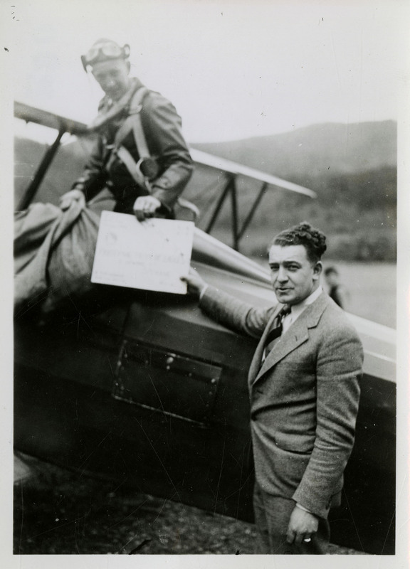Babe Peila, photographer for Tabor's Store, gives a Fraternal Order of Eagles certificate to pilot, George Nelson, who is standing up in the airplane and holding two bags.