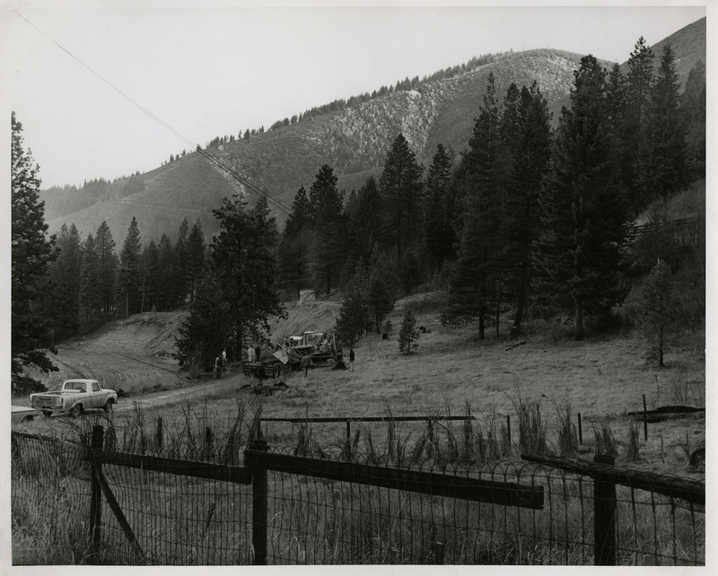 Logging equipment and two cars in the distance, several people surround the equipment, a man operates one of the machines. A fence is in the foreground, trees and mountains in the background.
