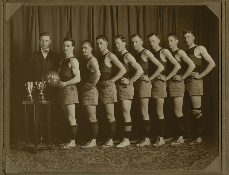The 1918-1919 Wallace High School basketball team pose with their coach. The first team member on the left holds a basketball with "W.H.S. '18-'19" written on it. Two trophies are displayed on a small table on the left. The photographer is Barnard. 