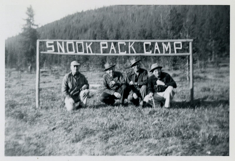 Four men squat underneath a sign reading, "Snook Pack Camp."
