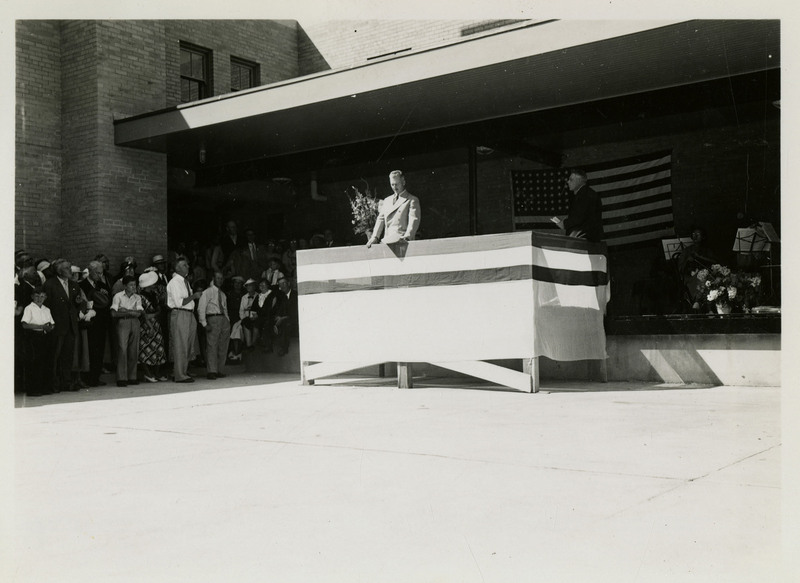 An unidentified man reads a speech at a dedication ceremony of the Wallace Post Office. People and children stand on the left. Musicians sit on stage with their instruments on the right. An American flag hangs on the building behind speaker and musicians.