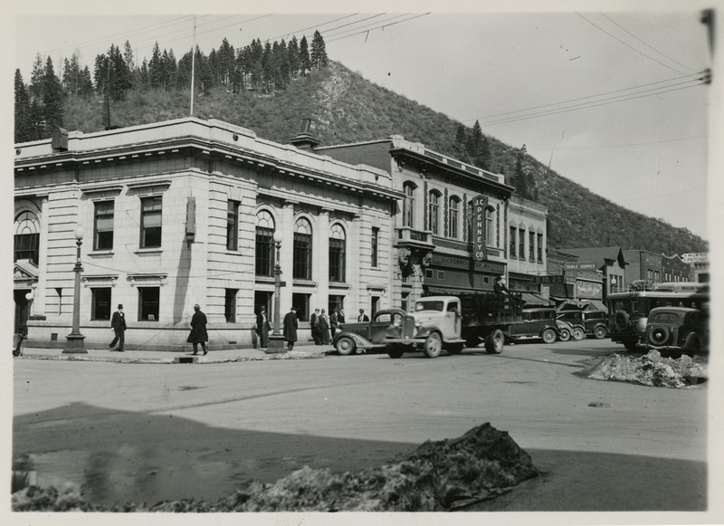 Street scene of Wallace, Idaho taken for Household Magazine. Several automobiles are parked or on the street. Men and women are walking on the sidewalk. Buildings including a J.C. Penney Co. line the street, with mountains and trees in the background. 