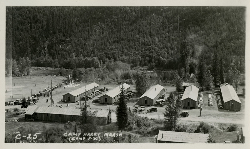 View of Camp Harry Marsh. Several buildings with automobiles parked next to it. A crowd of people surround a field on the left. Trees in the background. Caption on the photograph reads, "C-25 Camp Harry Marsh (Camp F-30).