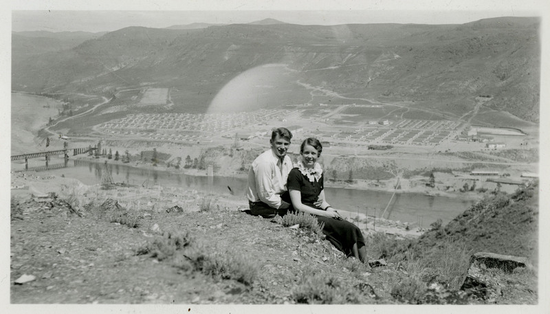 A man and woman pose in front of a view of the Grand Coulee Dam.