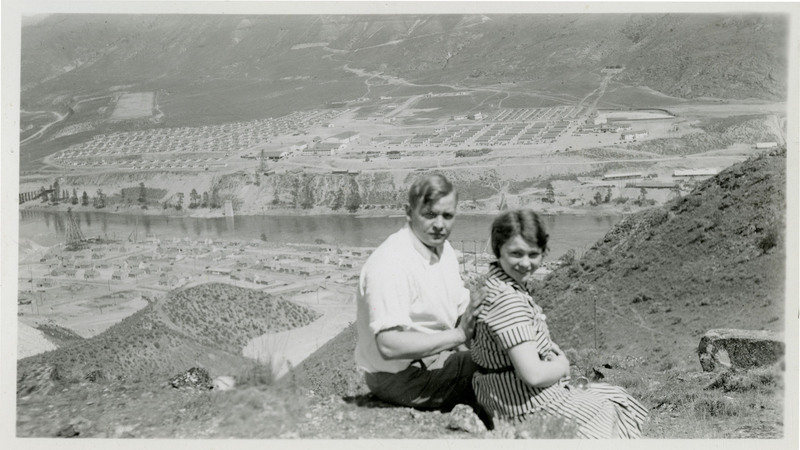 A man and woman pose in front of a view of the Grand Coulee Dam.