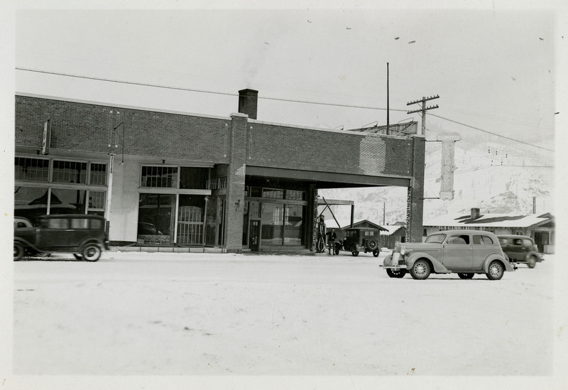 A view of the Yellowstone Trail Garage brick building. A man is using the gas pump. Several automobiles passing in front of the building. Snow covered mountains and trees in the background.
