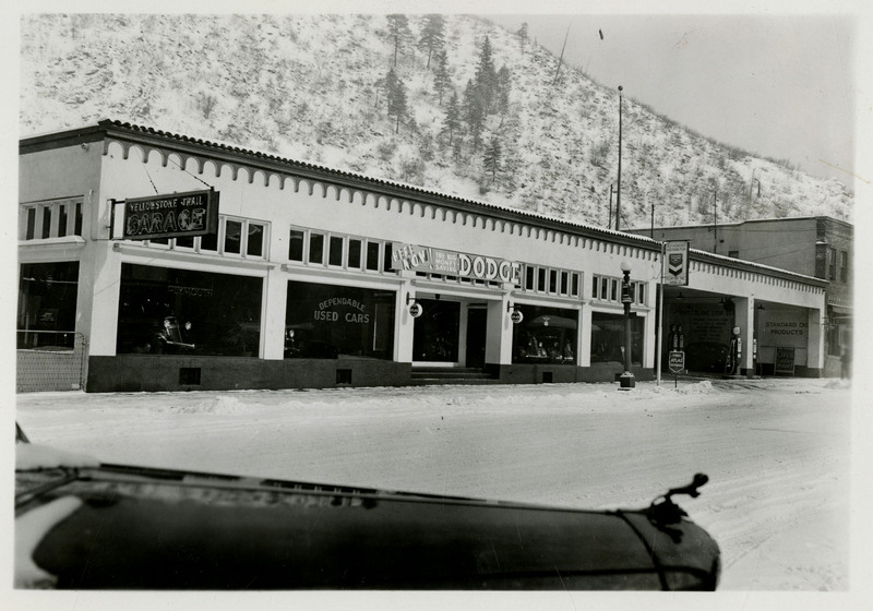 A view of the Yellowstone Trail Garage building. Part of an automobile is in the foreground, snowy mountains and trees in the background.