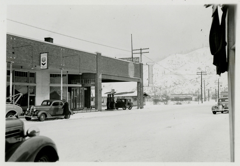 A view of the Yellowstone Trail Garage building. An automobile can be seen near the gas pumps. Other automobiles are parked or on the street. One man can be seen near a car.