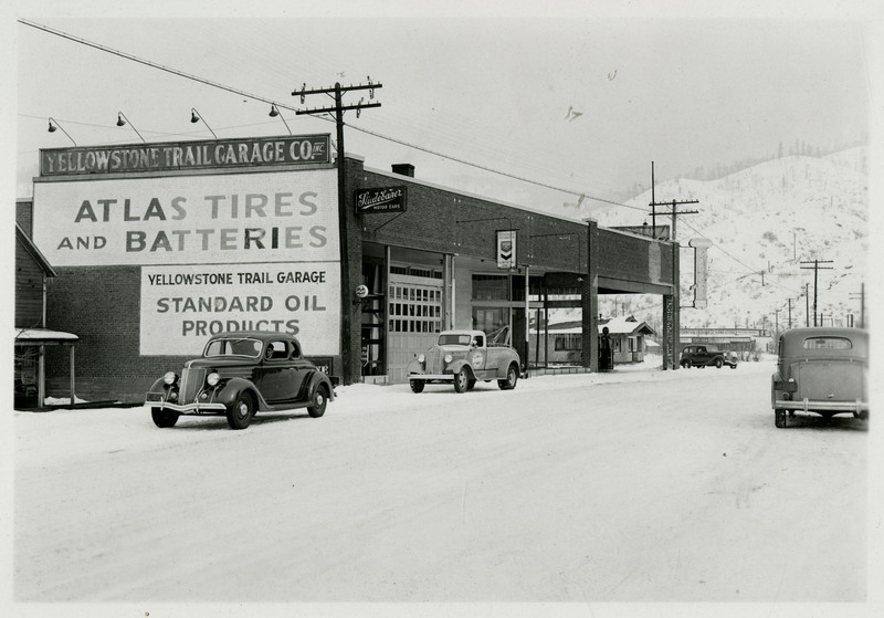 A view of the Yellowstone Trail Garage building. Large signs reading "Atlas Tires and Batteries" and "Yellowstone Trail Garage Standard Oil Products" can be seen on the side of the building. Several automobiles are on the snow-covered street.