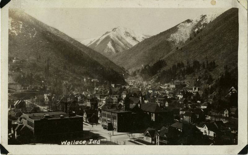 Postcard depicting a view of Wallace. Mountains and trees are in the background. Buildings and streets can be seen in the foreground.