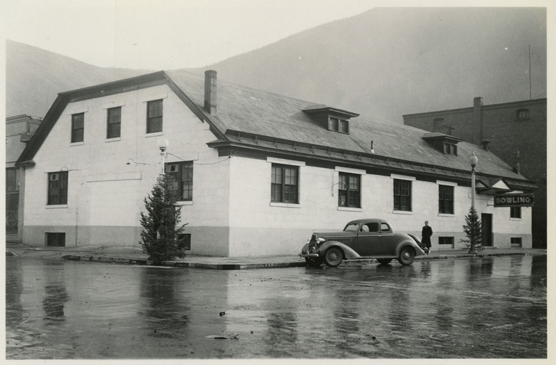 View of the bowling alley in Wallace. A man can be seen walking in front of the building. An automobile is parked on the street. On verson reads "Formerly the Sutherlands' Levery Stable Rom."