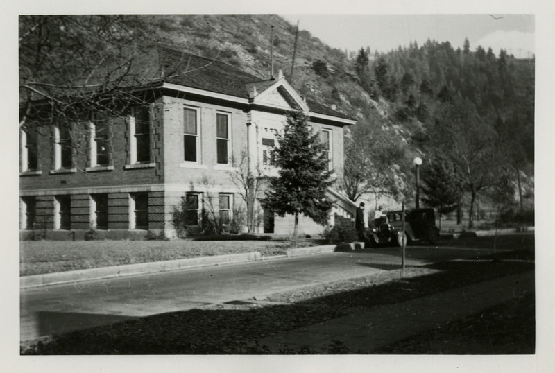 View of the Wallace Public Library. Two people stand near an automobile that is parked in front of the building. Trees and mountains are in the background.