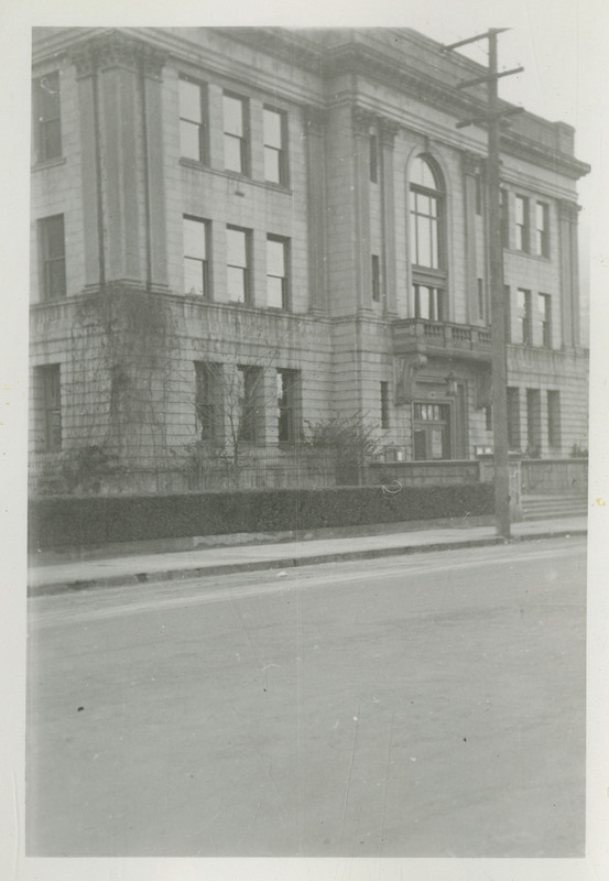 View of the Shoshone County Courthouse. A man can be seen walking in front of the building. An automobile is parked on the street. On verso reads "Formerly the Sutherlands' Levery Stable Rom."