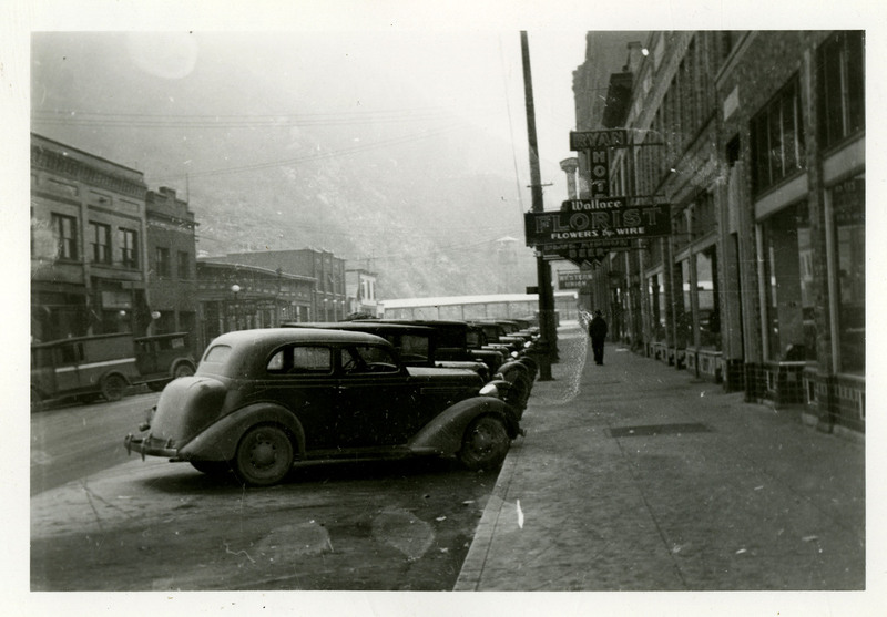 View of Cedar Street looking East from near 6th Street. Automobiles are parked along the sidewalk. A man is walking down the street in between the automobiles and the buildings. One of the building signs read "Wallace Florist Flowers by Wire."