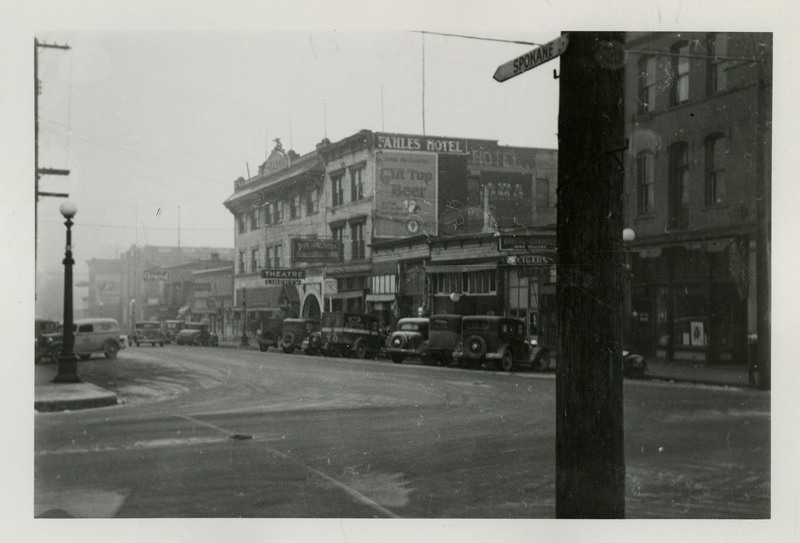 Street View of Wallace, Idaho. A street marker in the foreground reads "Spokane." Automobiles are parked alongside the street, near several storefronts, including a theatre, Fahles Hotel, and a cigar store.