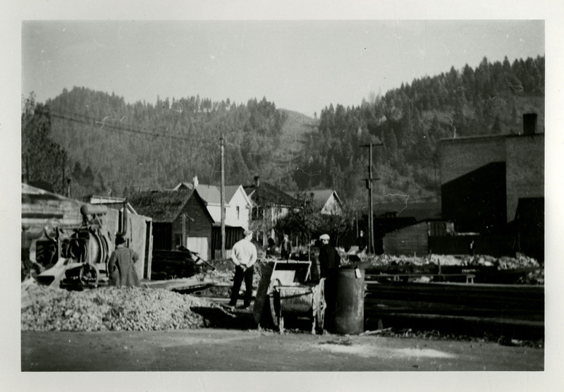 Unidentified building being constructed. Three men are standing near the construction site. Mountains and trees are in the background.