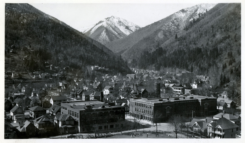 An overhead view of Wallace, Idaho. Buildings and a few people in a park are in the foreground, surrounded by trees and mountains in the background.