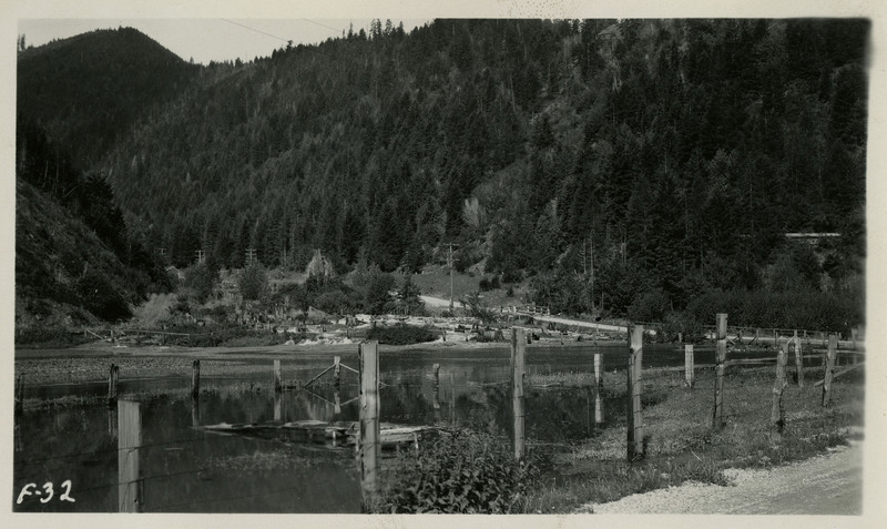A view of the mouth of Lake Gulch. A fence borders a lake in the foreground. Trees and a mountain landscape appear in the background.