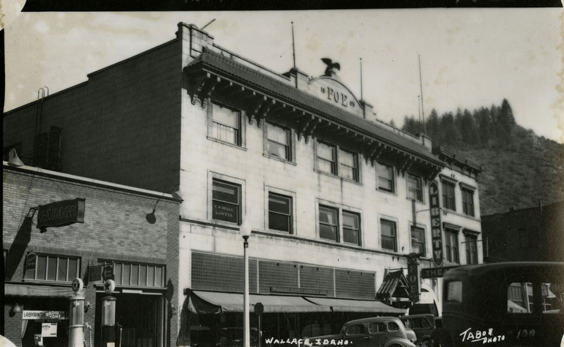 Postcard depicting a street view of the Fraternal Order of Eagles building in Wallace, Idaho. One store sign reads "Wallace Supply Company Ltd." Several automobiles are parked or driving by.