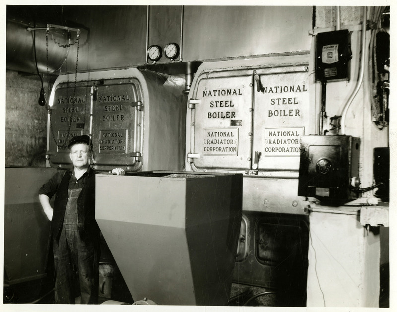 A man in overalls stands next to the Wallace High School boilers.  "National Steel Boiler" and "National Radiator Corporation" is inscribed several times on the boiler equipment.