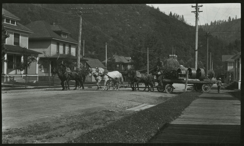 Two men operate a horse-drawn cart brings equipment to the Wallace light plant on First Street. A man can be seen on the right walking behind the cart.