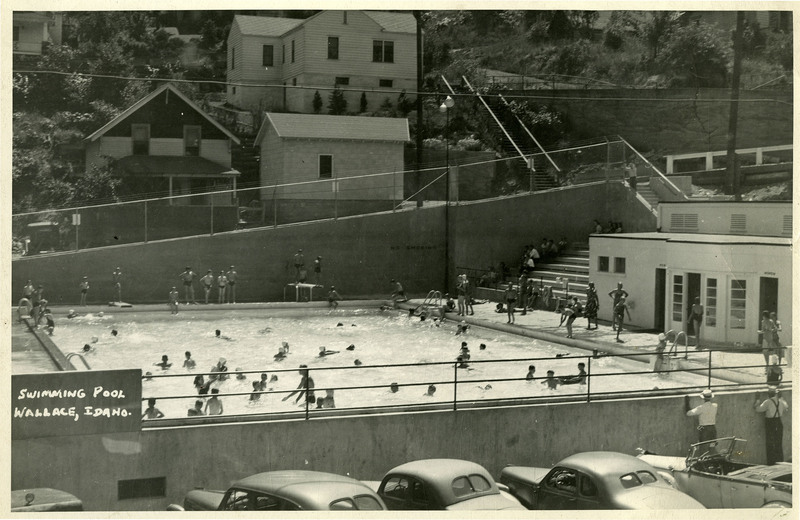 A view of the Wallace Swimming pool. People are in the pool, standing on the deck, or sitting on the bleachers. 