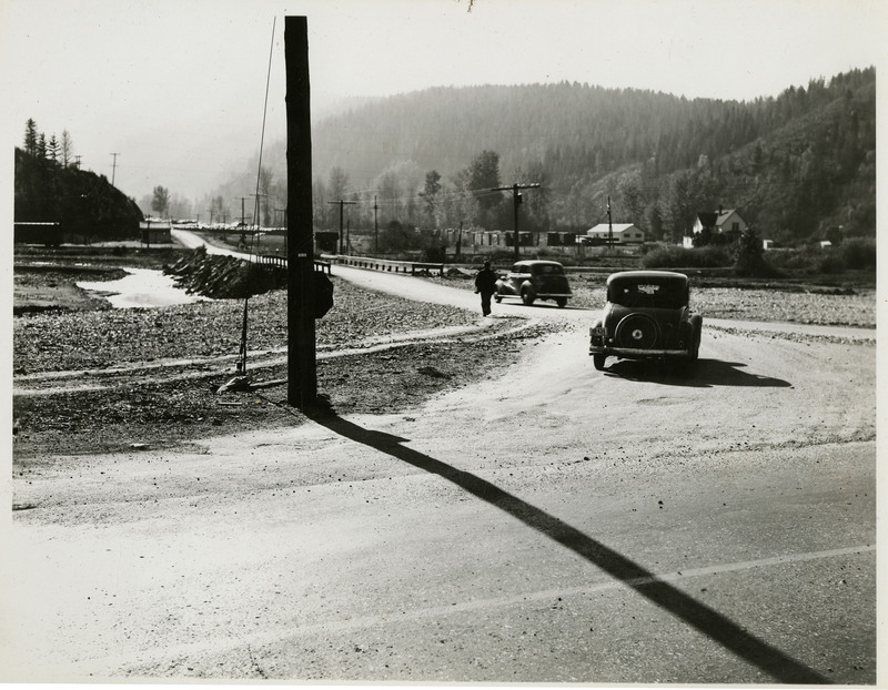 Two automobiles driving on an unidentified road. A man can be seen alongside the road. A few buildings and trees are in the background.