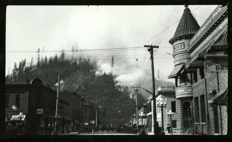Photo of 6th street of Wallace, Idaho, taken circa 1920. Several buildings, cars, and a few people are visible on the street, and in the background there is a tree covered hill. Two separate flag poles with the American flag are visible as well.