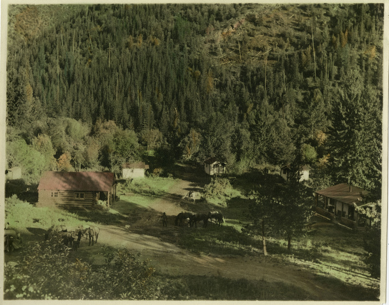 A view of an outdoor scene in an unidentified location. A few buildings, people, and horses can be seen.
