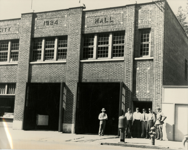 The City Hall building, which is inscribed with the text "CITY 1924 HALL". A man stands in front of the building, under a garage opening, holding a paper. Five other men wearing uniforms stand in front a smaller doorway.