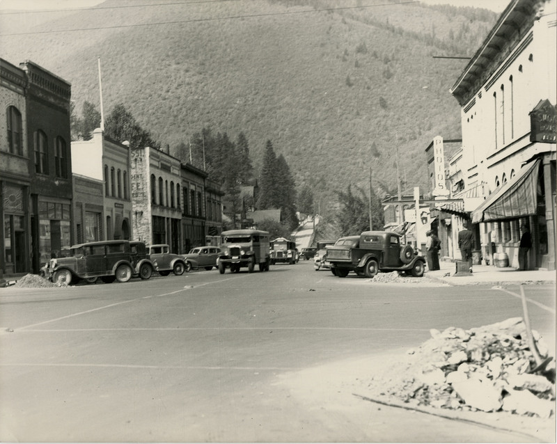 A street scene in Wallace, Idaho. Several automobiles are parked along the sidewalk and driving in the road. A few people can be seen on the right sidewalk.