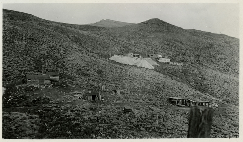 A photograph depicting a possible construction of a mine. A few buildings can be seen in the landscape. On verso reads "Big Mines from little mines must grow."