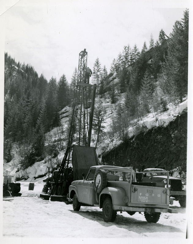A man stands on a crane which is next to a truck. Snow Covered landscape is in the background.