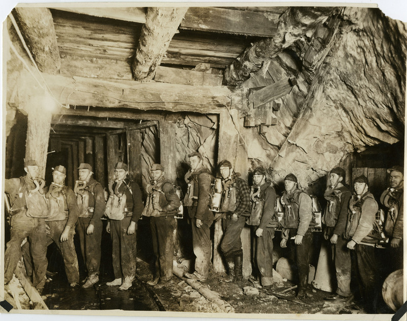 Miners pose wearing oxygen masks and tanks in a lit tunnel.