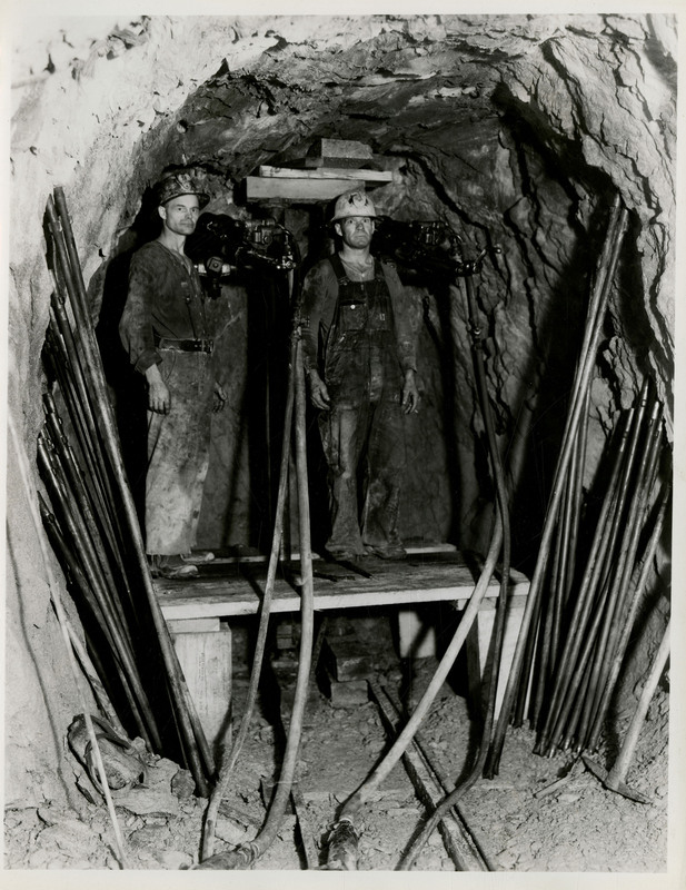 Two miners stand on a platform inside a mine.
