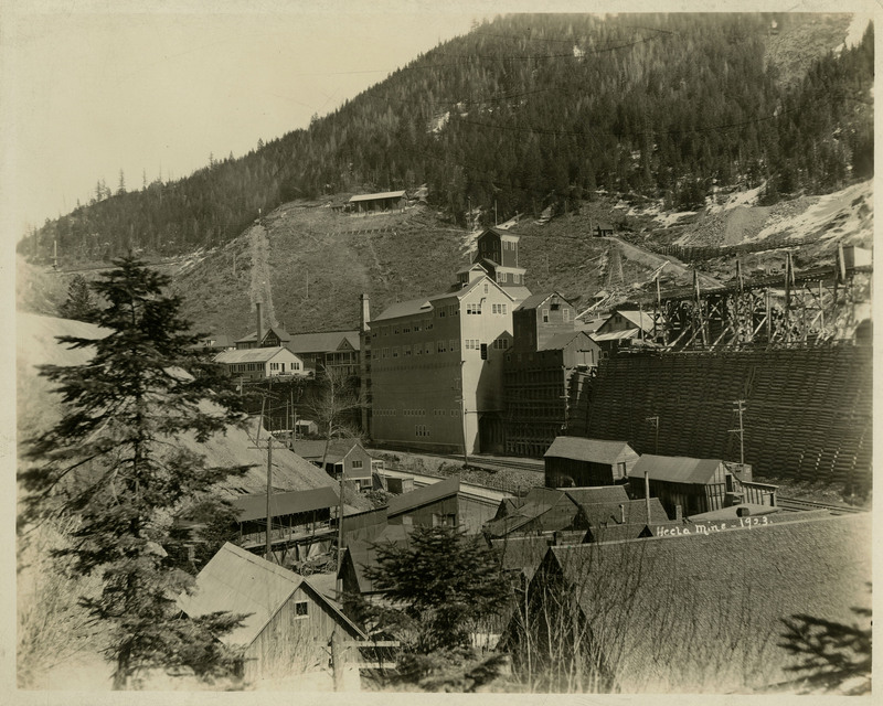 View of the Hecla Mine buildings with trees and mountains in the background.