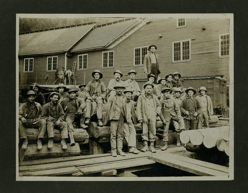 Hecla Mining Company miners sit on logs outside a building. Three men can be seen in the background near the building.