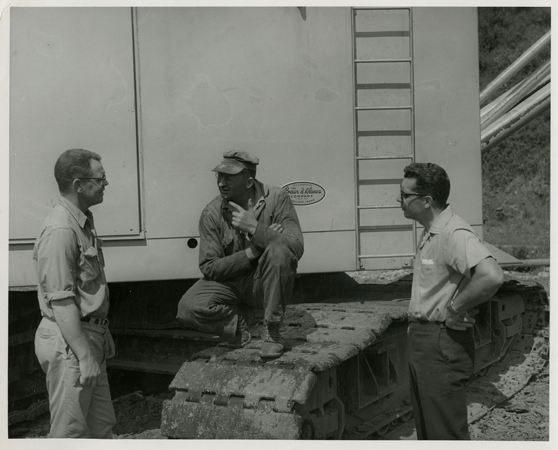 Three men discussing outside. Two of the men are standing, the man in the center is squatting on continuous tracks.