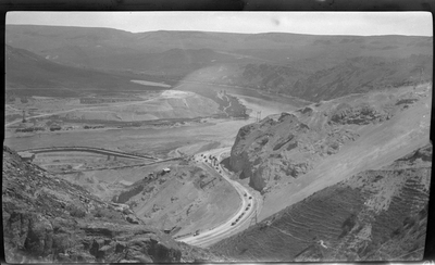 Several lines of cars are seen driving up the hill near Coulee Dam, with the river visible in the background.