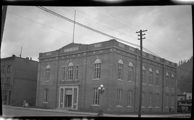 Photo of the Benevolent and Protective Order of Elks building in what is likely Wallace, Idaho. There are two people walking down the road in front of the building, one of which is pushing a baby carriage.