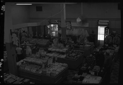 Interior photo of the J. C. Penney Company store. Various clothing items are on display for sale including shirts, skirts, lingerie, and pants. At least four people can be seen shopping.
