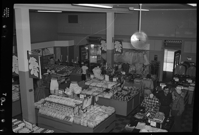 Interior photo of the J. C. Penney Company store. Various clothing items are on display for sale including shirts, skirts, lingerie, and pants. At least four people can be seen shopping.
