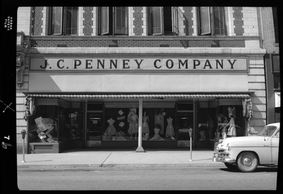 Photo of the storefront of the J. C. Penney Company building. There are several mannequins displaying various clothing items in the front windows, and a car parked on the side of the street next to the building.