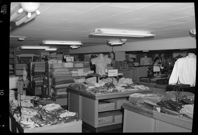 Interior photo of the J. C. Penney Company store. There are several displays of various clothing pictured, including shirts, pants, and dresses. A woman can be seen shopping in the background.