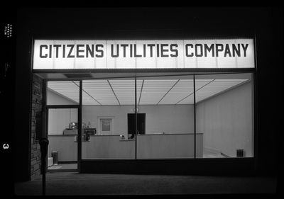The storefront of Citizens Utility Company. The light of the sign above the entrance and the interior lights are on.