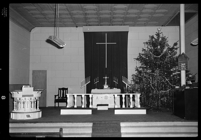 Photo of an altar of an unspecified Lutheran Church in Mullan, Idaho. The altar has a large Christmas tree in one corner that is decorated. The altar itself stands in the middle of the area with candle sticks around it.