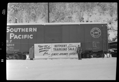 Photo of two men standing on either side of a banner advertisement for Elmer's Appliance Center that is hung across a Southern Pacific train car. The banner reads, "This car of 1960 Hotpoint Appliances for Elmer's Appliance Center; Hotpoint 55th Anniversary Trainload Sale!" There is a heavy layer of snow covering the ground and the top of the train car.