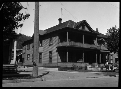 Photo of a home in Wallace, Idaho taken for Household Magazine. The house is two stories with an alley on the side of it, and a balcony for the second story. There are a few people standing on the grass out front.